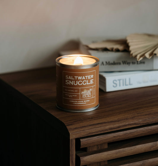 warm glowing Saltwater Snuggle soy candle on a retro wooden side table in a cozy Cornish cottage with coastal living books in the background— eco soy candle for relaxing home vibes