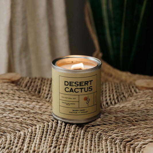 Lit Desert Cactus soy candle in an earthy green tin, glowing warmly on a natural woven stool with a desert plant behind it — an eco-friendly, vegan candle perfect for nature lovers and boho interiors.