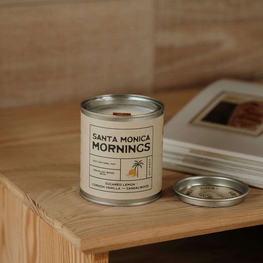 Santa Monica Mornings soy candle, with a wooden wick on light wood bedside table with surf shack book open in background, capturing coastal surf lifestyle and natural candle vibes.