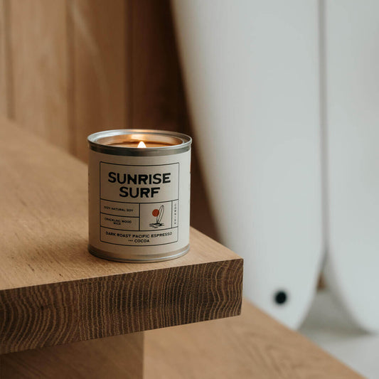 Sunrise Surf espresso soy candle lit on a wooden step in a Californian-style home with a white surfboard in the background, capturing laidback surf vibe