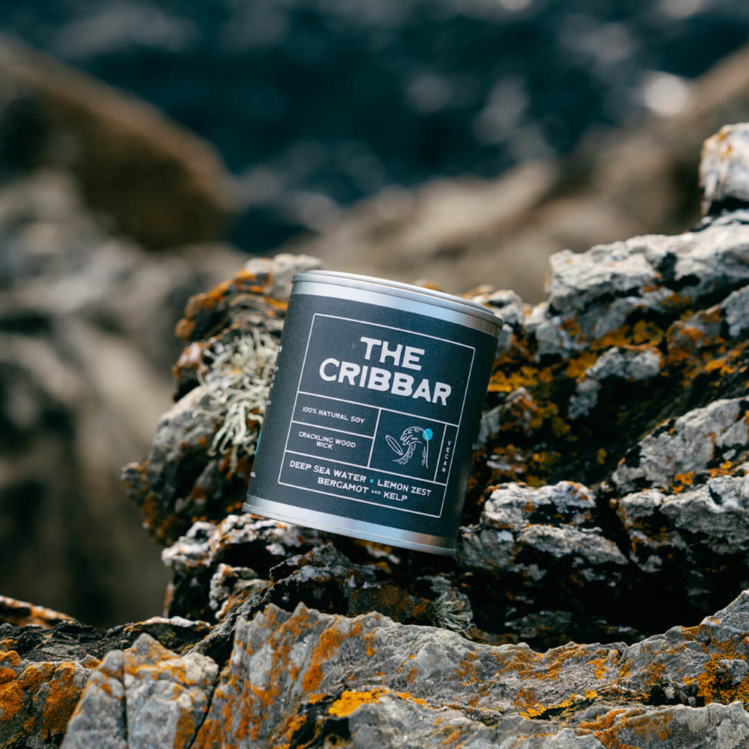 The Cribbar limited edition ocean-scented soy candle by Kernowfornia Soul, clean burning and eco friendly, in black label tin on Newquay’s rugged coastal rocks with deep blue sea—perfect surfer gift from Cornwall