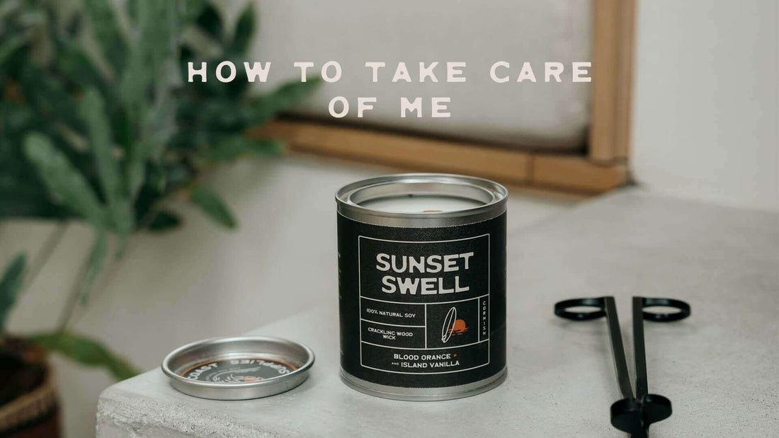 "Eco-friendly soy candle with crackling wood wick in black 'Sunset Swell' tin, placed on a minimalist concrete surface with wick trimmer and tropical plant in background – scented with blood orange and island vanilla."