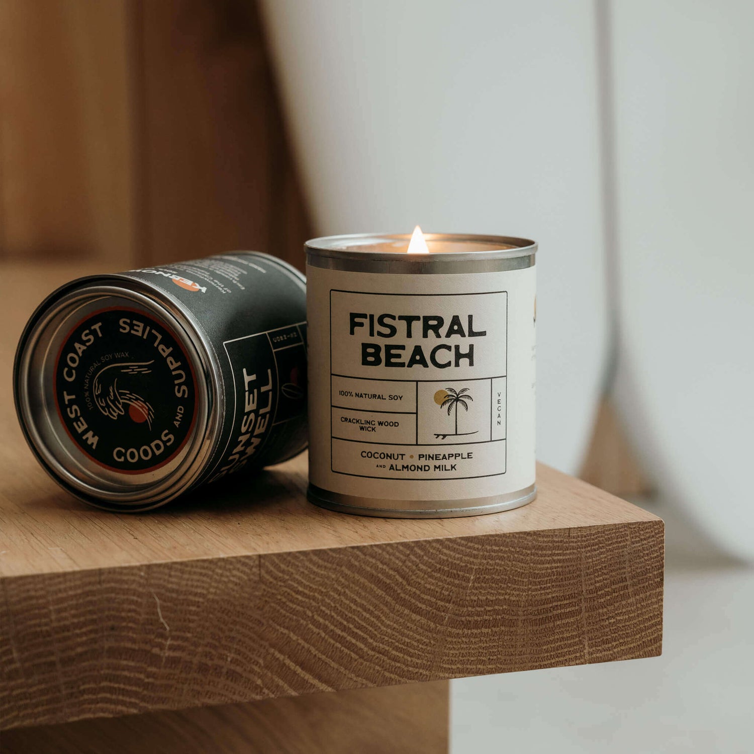 Black eco soy candle tin with wave logo next to lit Fistral Beach soy candle with wooden wick on a step, surfboard in background, coastal surf lifestyle vibe.