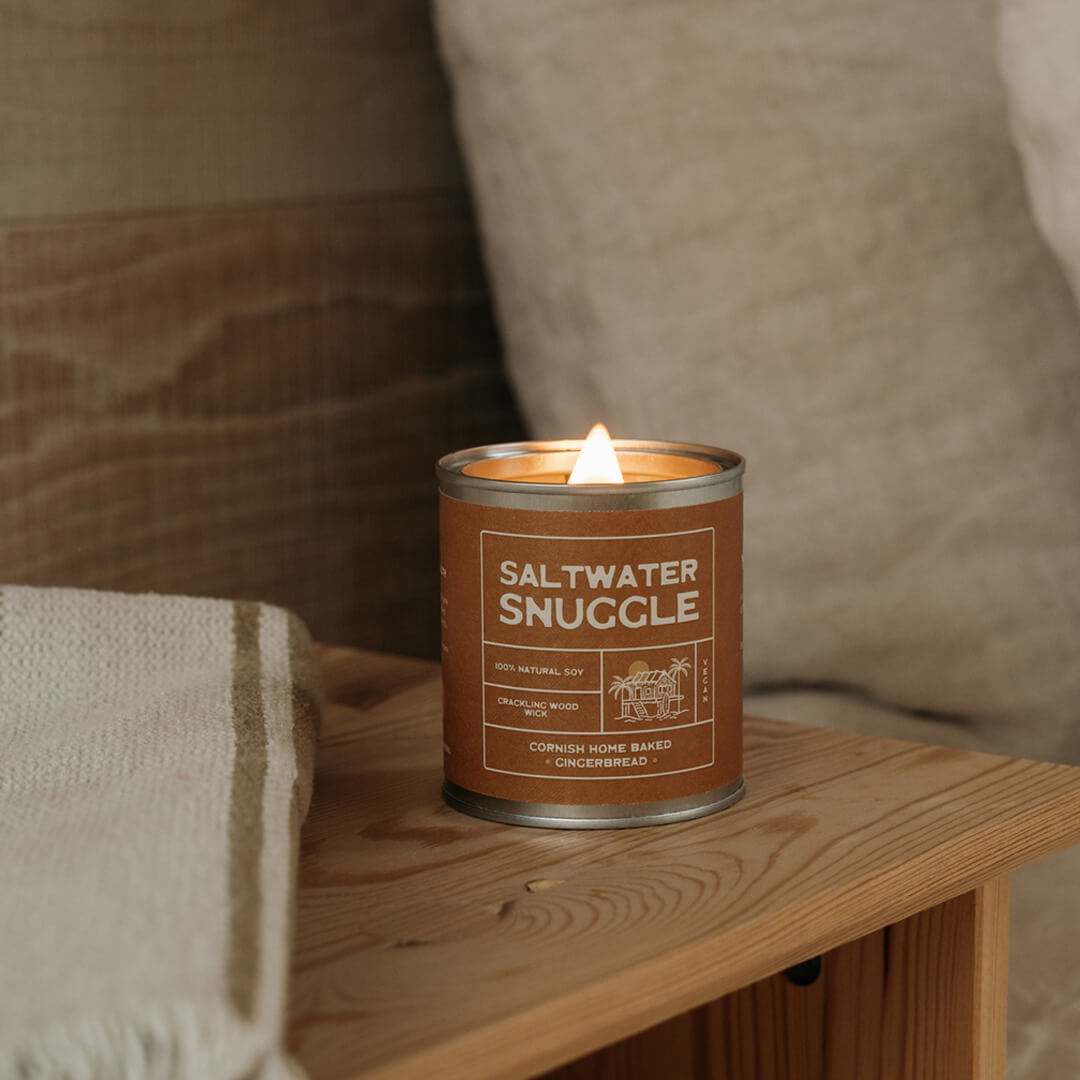 Saltwater Snuggle gingerbread soy candle on a wooden side table in a cozy Cornish cottage bedroom with natural linen bedding — eco soy candle for relaxing home vibes