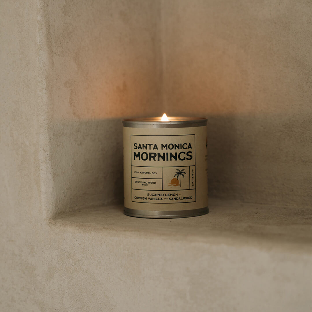 Lit Santa Monica Mornings soy candle on a micro cement shelf in a Californian-inspired cafe, featuring a clean-burning natural soy wax candle with wooden wick and relaxed coastal lifestyle vibes.